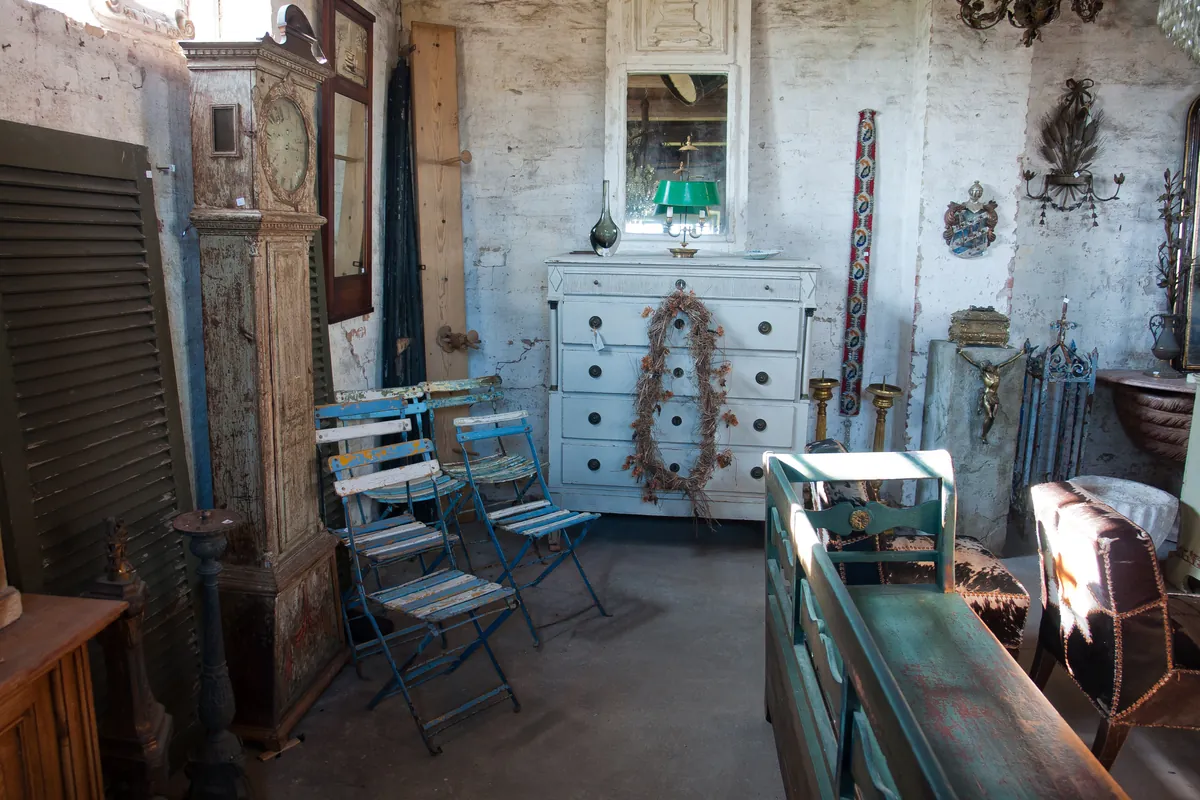 Grenier rempli de mobilier ancien : horloge comtoise, commode peinte, chaises de jardin pliantes